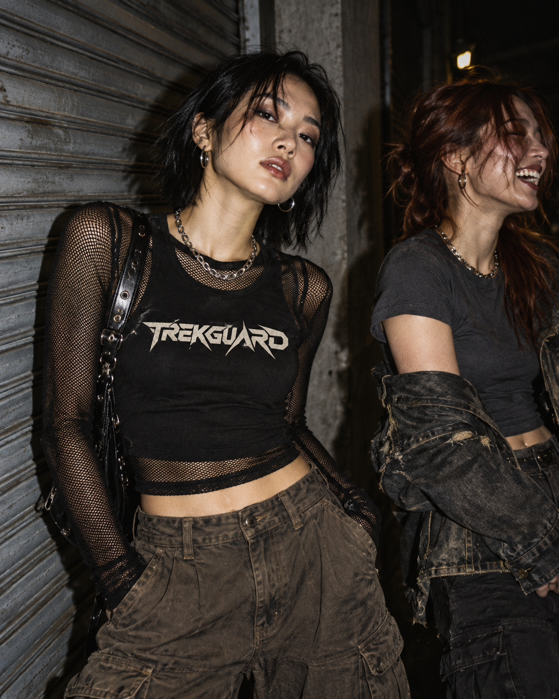 Two women in dark streetwear beside an industrial shutter, one wearing a TrekGuard top